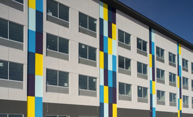 Exterior shot of Tru by Hilton hotel in Chicopee, MA. Predominantly grey with blue, purple, and yellow rectangular accents.